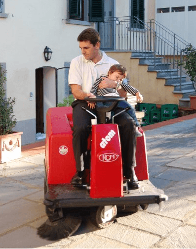 Un uomo pulisce il cortile in pietra con una macchina mentre tiene in braccio una bambina sorridente.