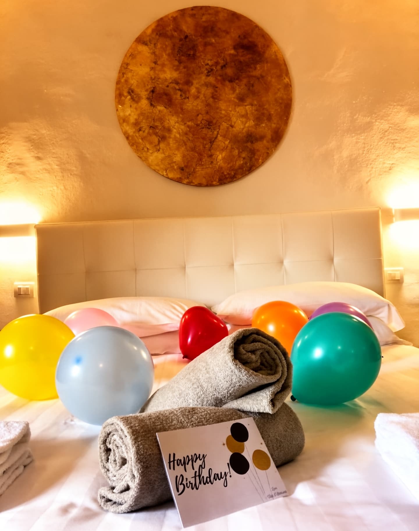 Interno di una camera elegante, decorata con palloncini colorati e asciugamani arrotolati, un messaggio di auguri di compleanno.