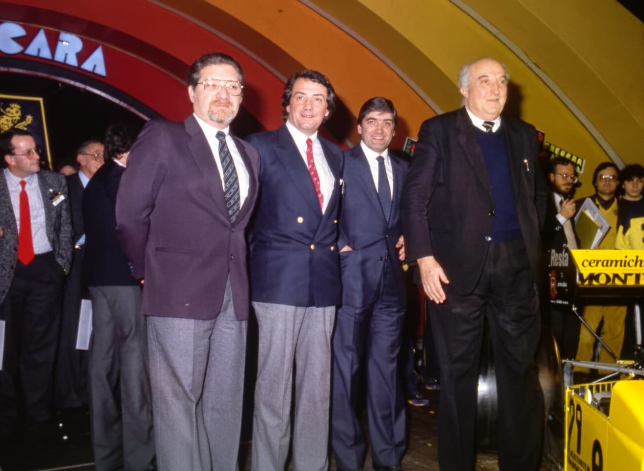 Quattro uomini in abiti formali sorridono in un ambiente con decorazioni vivaci, richiamando la storia della Formula 1.